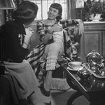 "Designer Anne Fogarty, dressed in plaid taffeta outfit with white lace pantaloons while enjoying a visit from a friend in her apartment."